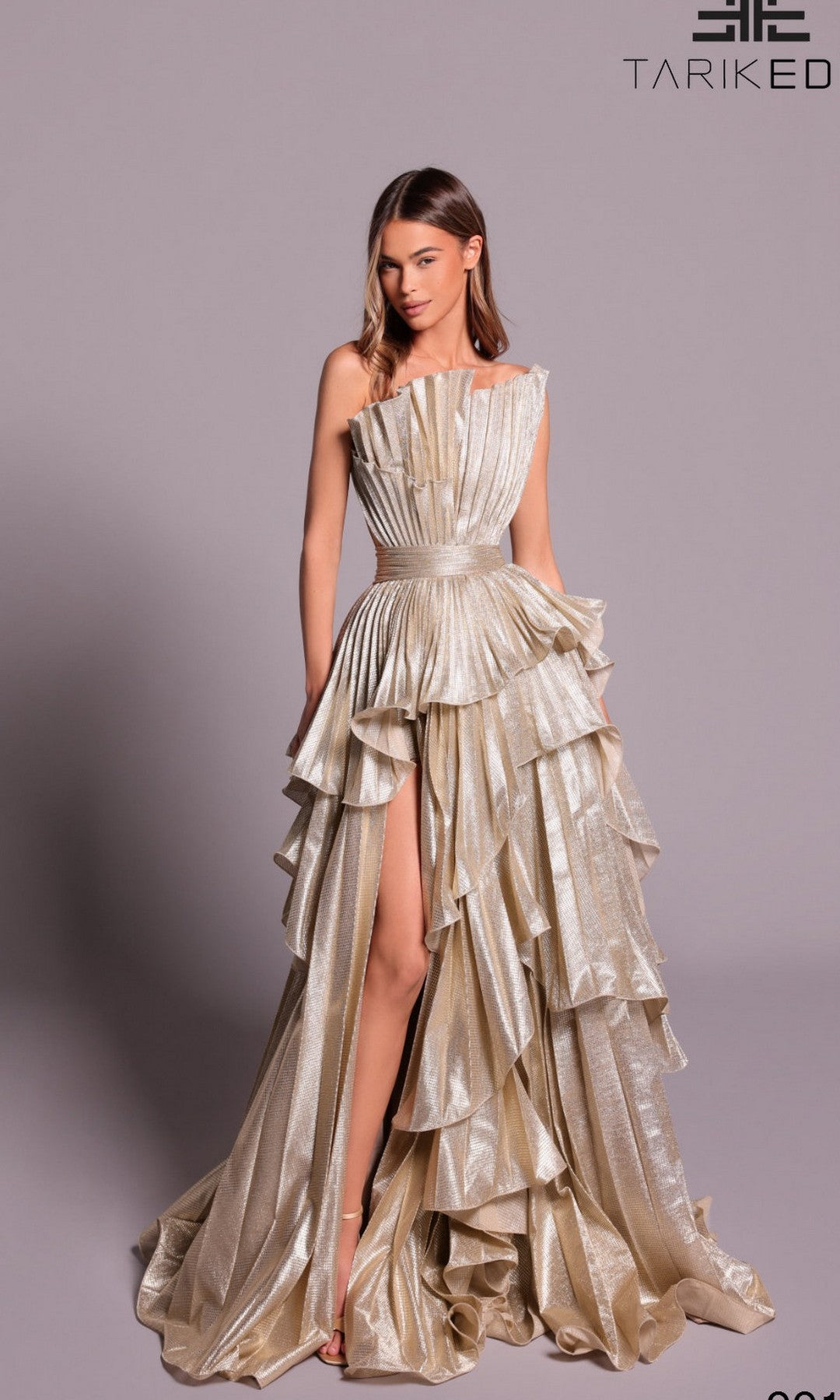 A woman models the Tarik Ediz 99148 long formal dress in shimmering gold, featuring a one-shoulder design, high slit, structured waist, and flowing layers—perfect for a red-carpet look against a plain backdrop.